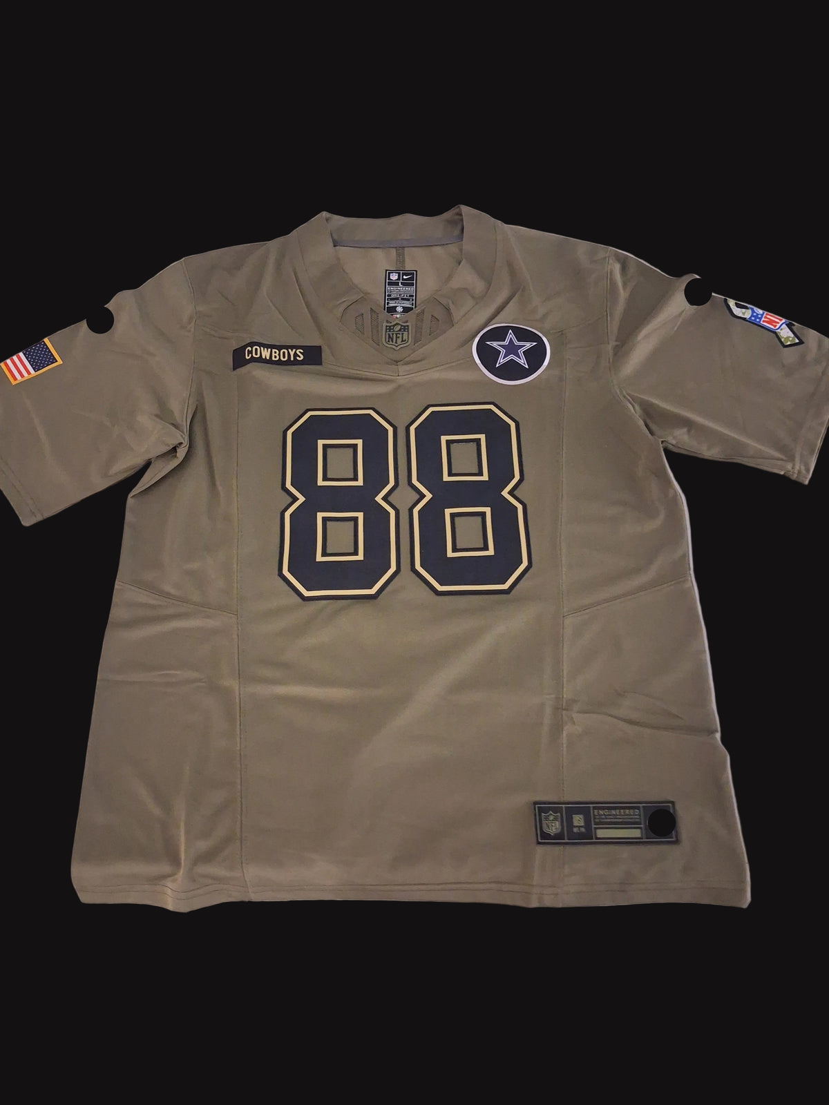 A Dallas Cowboys Nike football jersey in olive drab with the number 88 in black and gold on the front, laid flat on a gray textured surface.
